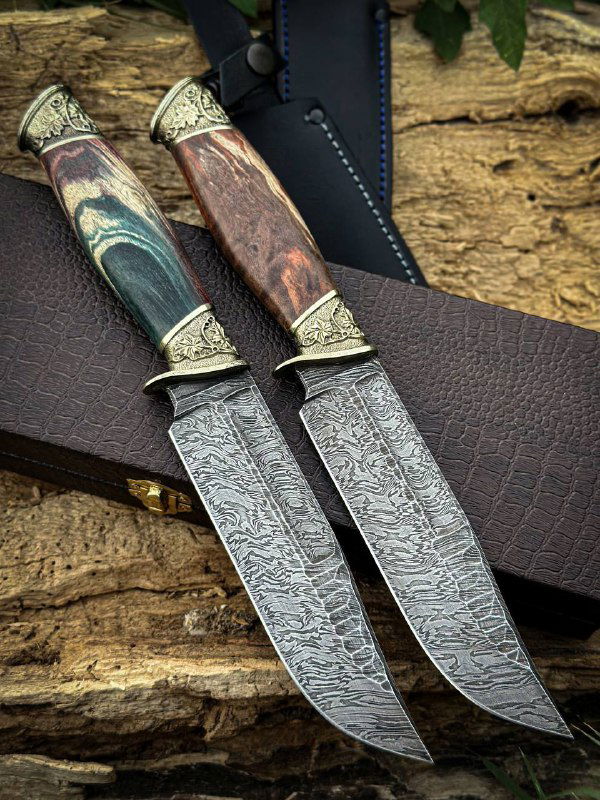 Крупный план двух дамасских клинков с волнообразным узором, видны мельхиоровые гарды и контраст древесины рукоятей.