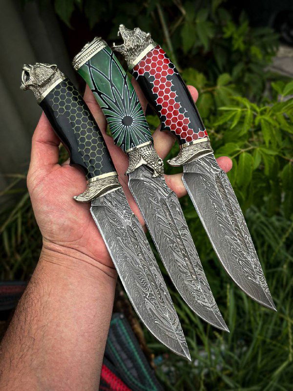 Три ножа в руке мастера: видно рисунок дамаска на клинках и текстуру акриловых рукоятей с мельхиоровыми гардами, крупный план.