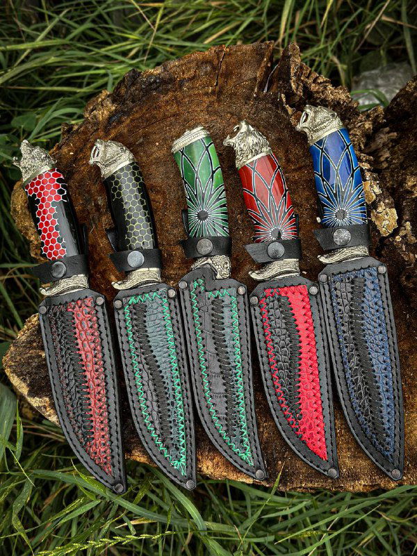 Четыре ножа с ножнами, расположенные на коре и траве; вид с ножнами внизу, различимые цвета вставок в швах ножен и техника пошива кожаных чехлов.