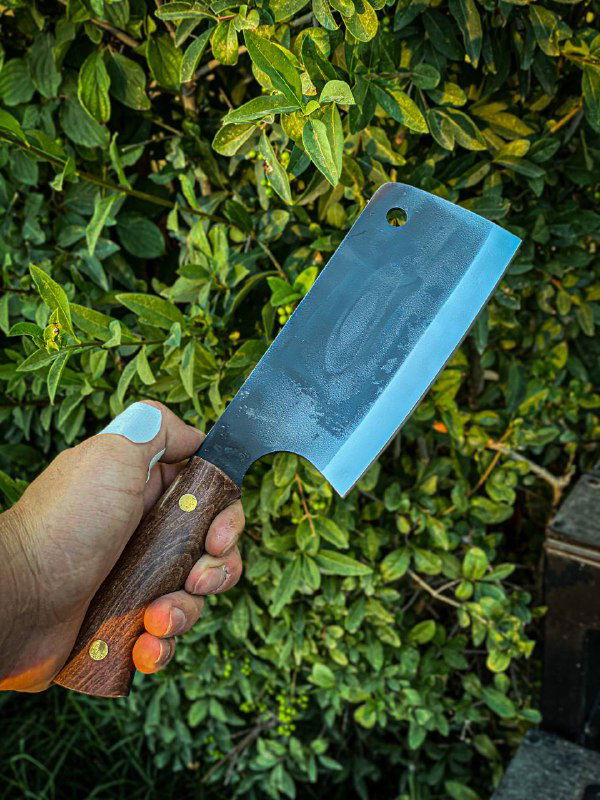 Крупный план одной тяпки из стали 95х18 в руке на фоне зелёных кустов, видно рисунок металла и фактуру рукояти из кавказского ореха.