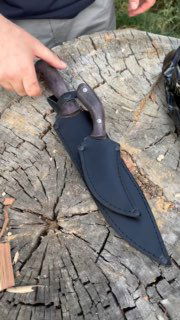 Скоторез в чёрных кожаных ножнах на пне: видна форма ножен, рукоять из орехового дерева и стальной клинок с характерной фактурой.