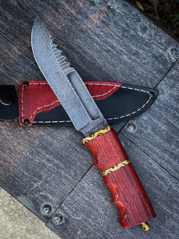 Крупный план ножа и кожаных ножен: текстура дамасской стали видна на клинке, чехол с красной прострочкой лежит на столе.