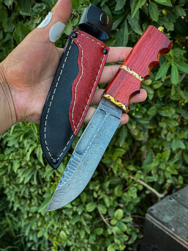 Нож в руке на фоне зелёной растительности: видно резную форму рукояти из карельской берёзы и прикреплённые ножны.