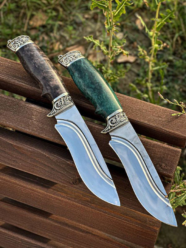 Два ножа с изогнутыми клинками и стабилизированными рукоятями тёмного и зелёного оттенка, крупный план на гарды с орнаментом мельхиора.