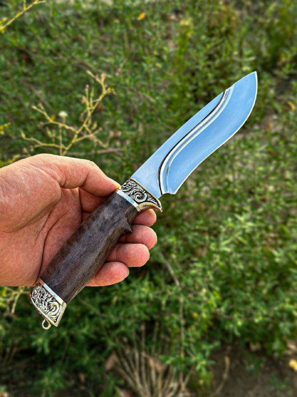 Рука держит нож с изогнутым клинком и зелёной стабилизированной рукоятью на фоне природы, видно полированное лезвие и декоративную гарду.