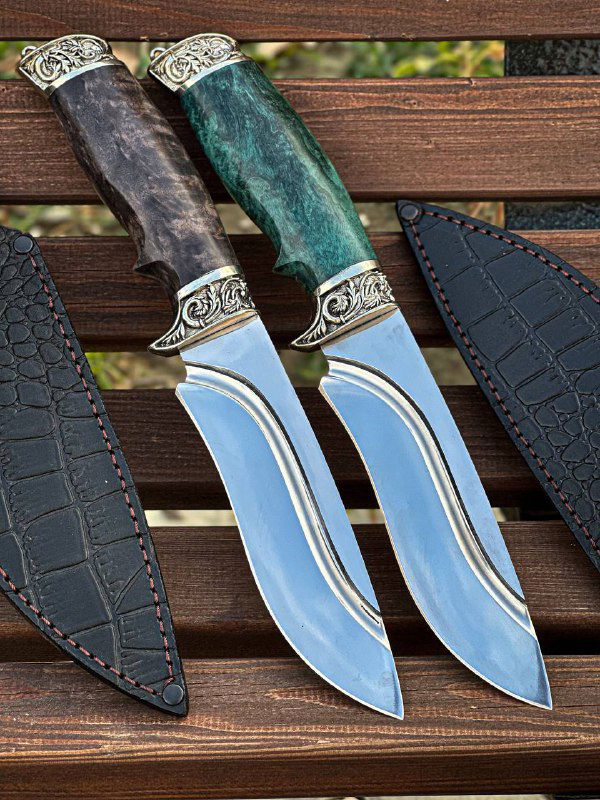 Два ножа рядом на деревянной поверхности в кожаных ножнах, видно форму клинков и совпадающие декоративные элементы на гардах и навершиях.