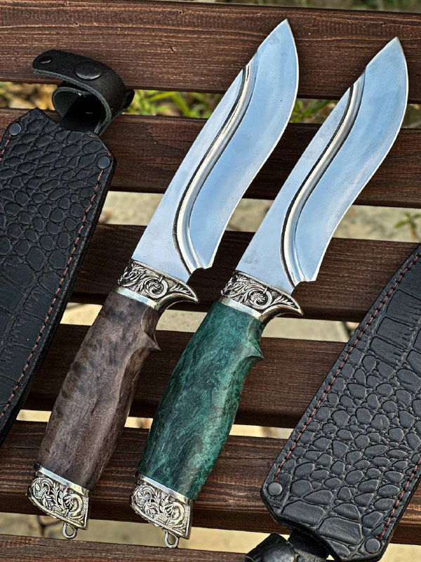 Два ножа в ножнах на столе, акцент на лезвии и форме клинка, видно швы и текстуру кожаных ножен на коричневом фоне.