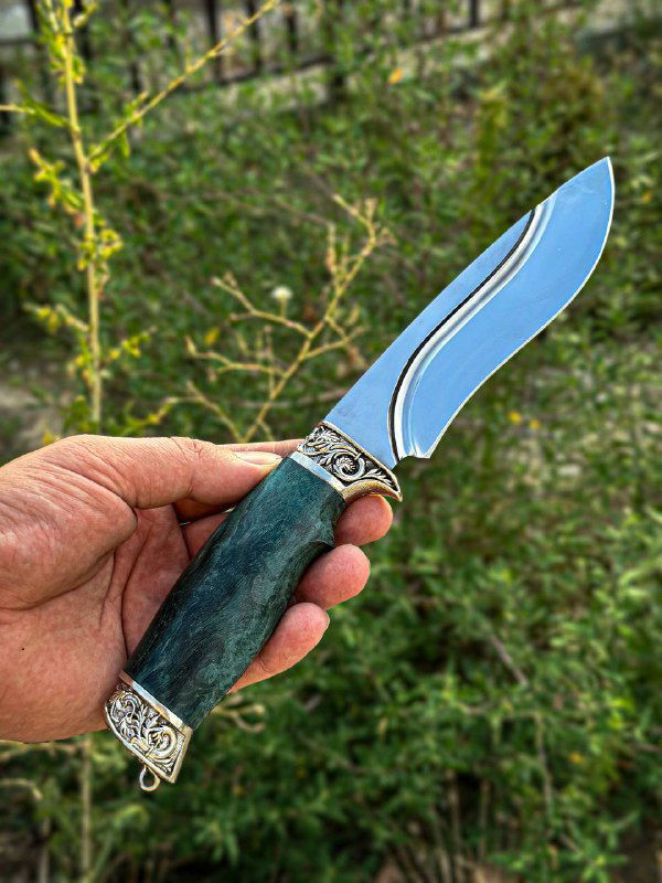 Крупный снимок ножа в руке на фоне зелёной растительности, показаны форма клинка, полировка и зелёная стабилизированная рукоять.