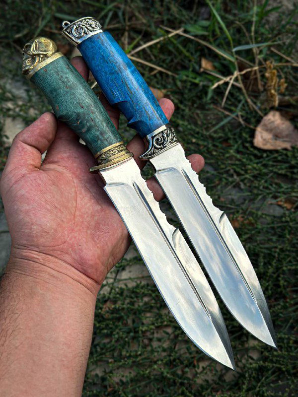 Два ножа в руке, вид снизу на клинки и навершия, освещение подчёркивает полировку и контраст между металлом и цветными рукоятями.