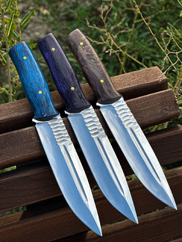 Три ножа рядом на скамейке, акцент на яркие стабилизированные рукояти (синий, фиолетовый, коричневый) и рифление у основания клинка.