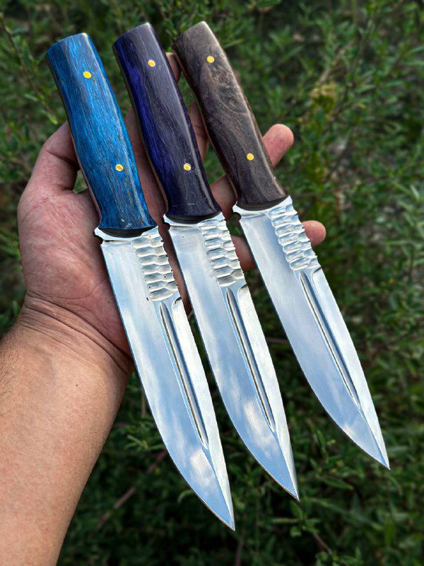 Руки держат три ножа в поле: крупный план клинков со шлифовкой и разноцветных рукоятей из карельской берёзы на фоне травы.