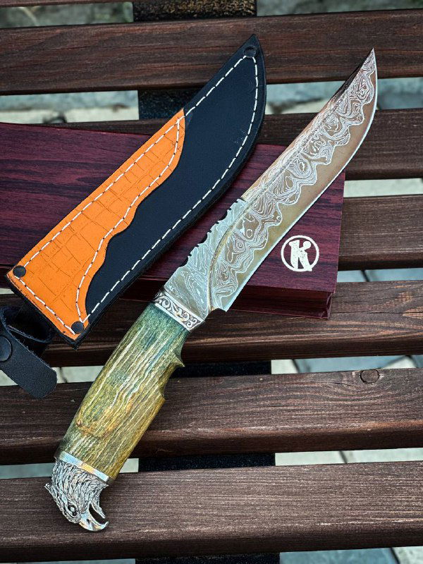 Вид ножа и ножен сверху: видно рисунок дамаска, кожаные ножны с оранжевой вставкой и текстуру карельской берёзы на рукояти.