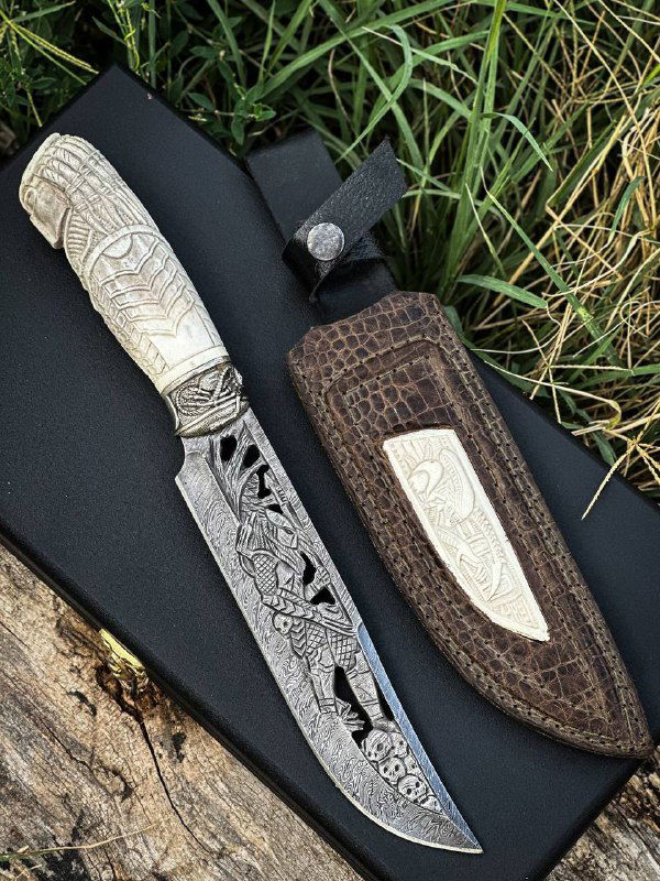 Крупный план ножа «ВОИН» в ножнах: украшенный клинок из дамаска с вырезной декоративной резьбой и кожаные ножны с вставкой из рога лося, фон — ткань и трава.