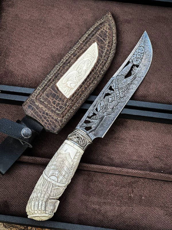 Полный вид ножа «ВОИН» рядом с ножнами: клинок дамаск, резная рукоять из рога лося и кожаный футляр лежат на коричневом фоне на природе.