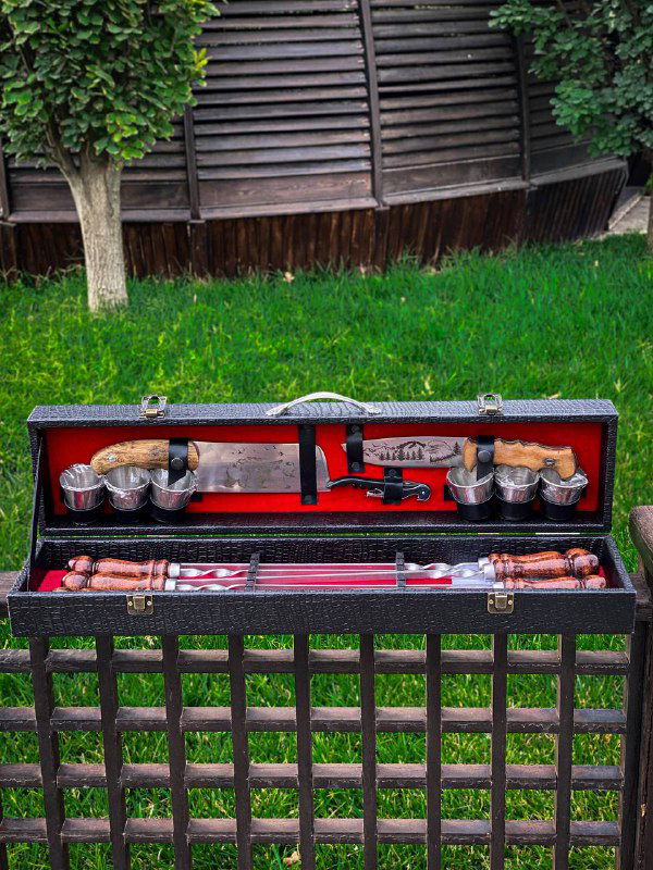 Фотография раскрытого кожаного кейса с шашлычным набором: видна тяпка, шампуры с деревянными ручками и набор металлических стопок.