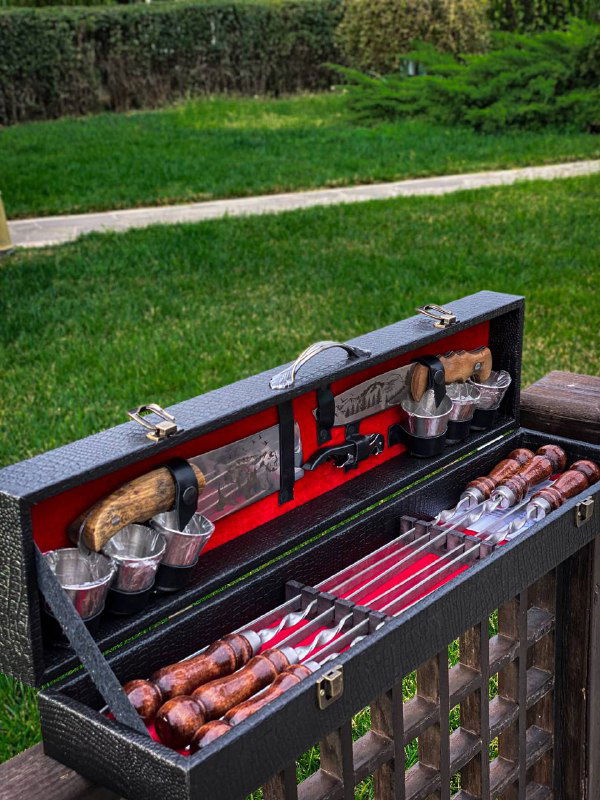 Шашлычный набор в открытом кейсе на зелёной лужайке: несколько шампуров, разделочный нож и металлические стаканы в отсеках кейса.
