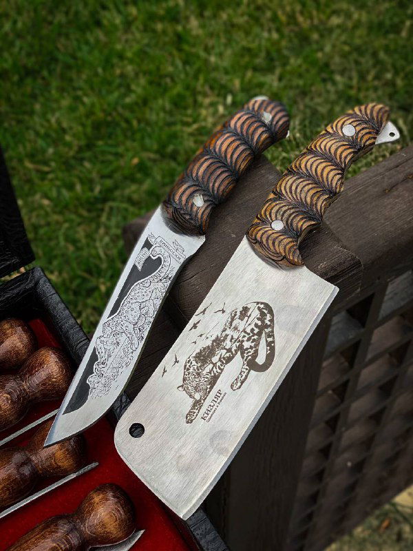 Крупный план двух кухонных клинков с гравировкой и декоративной отделкой рукоятей, видно структуру и маркировку стали на фоне ограды и травы.