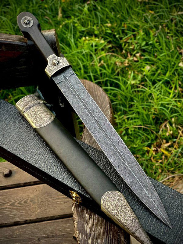 Крупный план боевого кинжала: дамасская текстура лезвия, рукоять из чёрного граба и кожаные ножны на деревянной поверхности.