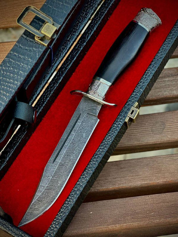 Крупный план ножа «Разведчик»: видно дамасскую текстуру клинка, мельхиоровые гарды и гладкую чёрную рукоять из граба, на красном фоне футляра.