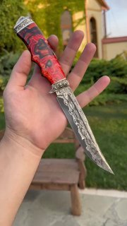 Нож в ладони для оценки размера: видна красно-чёрная акриловая рукоять, дамасковый узор на лезвии и мельхиоровые гарды, фон — сад и дом.