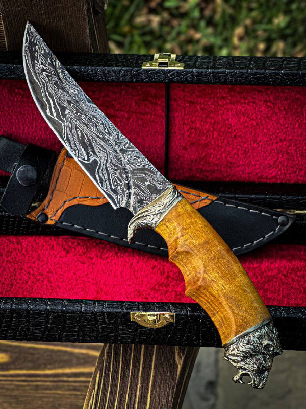Крупный план ножа на открытом футляре: заметен сложный дамасковый рисунок клинка, фактура карельской берёзы рукояти и металлические декоративные элементы.