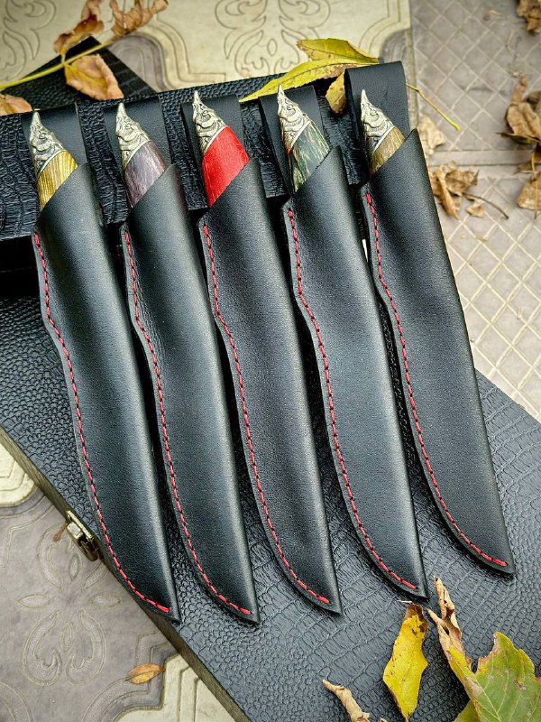 Пять чёрных кожаных ножен с красной строчкой для ножей «Осётр», выложенные в ряд на сером фоне с осенними листьями.
