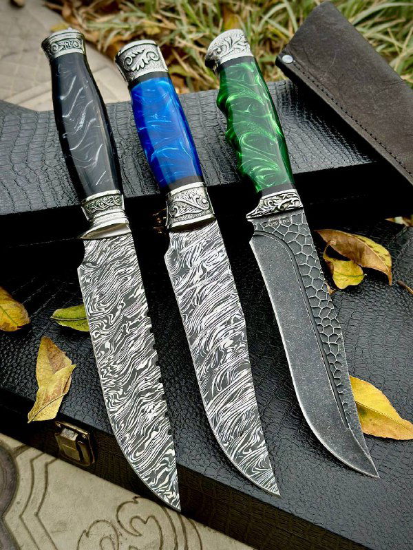 Три лезвия вблизи на темном фоне: выраженный дамасский узор, полированная поверхность и яркие акриловые рукояти — чёрная, синяя и зелёная.