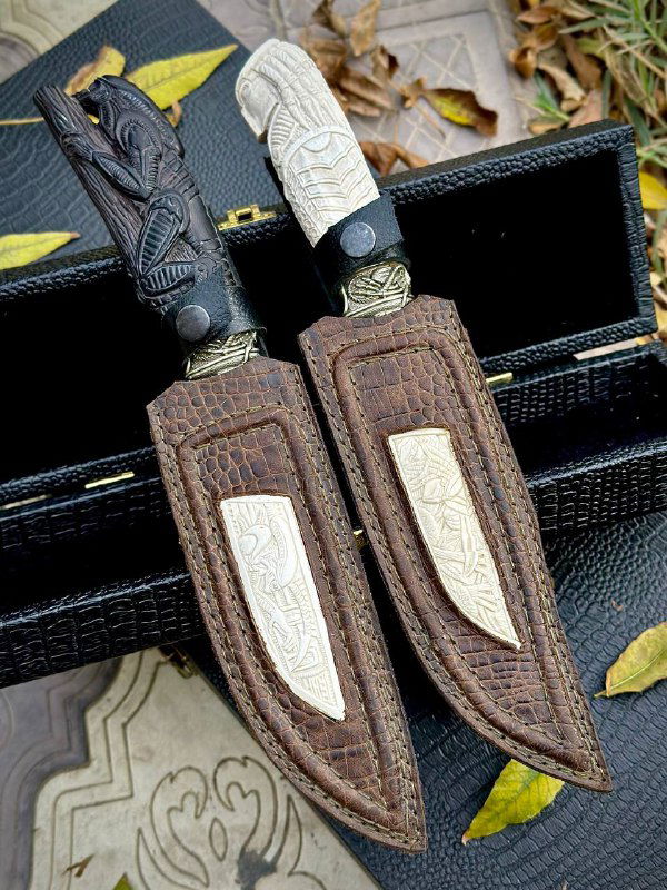 Кожаные ножны с вставкой из рога лося и аккуратной отделкой, рядом лежат два ножа с резьбой и мельхиоровыми элементами, вид сбоку.