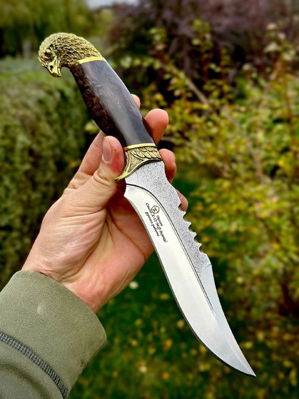 Нож «Орлан» в руке с тёмной рукоятью: крупный план латунной гарды, зубчатая спинка клинка и маркировка на лезвии из стали Х12мф.