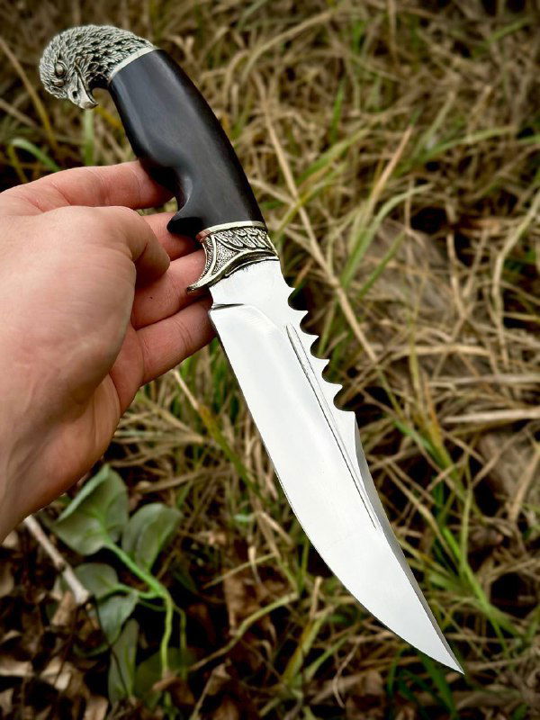 Боковой ракурс ножа «Орлан»: профиль клинка с насечками, декоративная мельхиоровая гарда и тёмная рукоять из граба на траве.