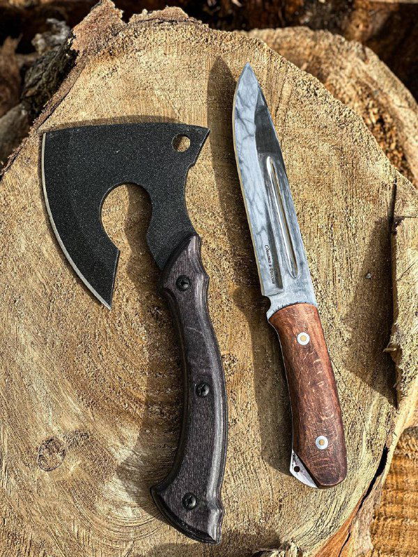 Крупный план ножа и топора на пне: заметна фактура стали клинка ножа и текстура рукоятей, видимая деталь отделки и поверхность металла.