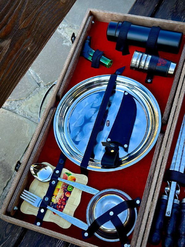 Крупный план тарелки, туристического ножа и нескольких аксессуаров набора для шашлыка на красной подкладке кейса.