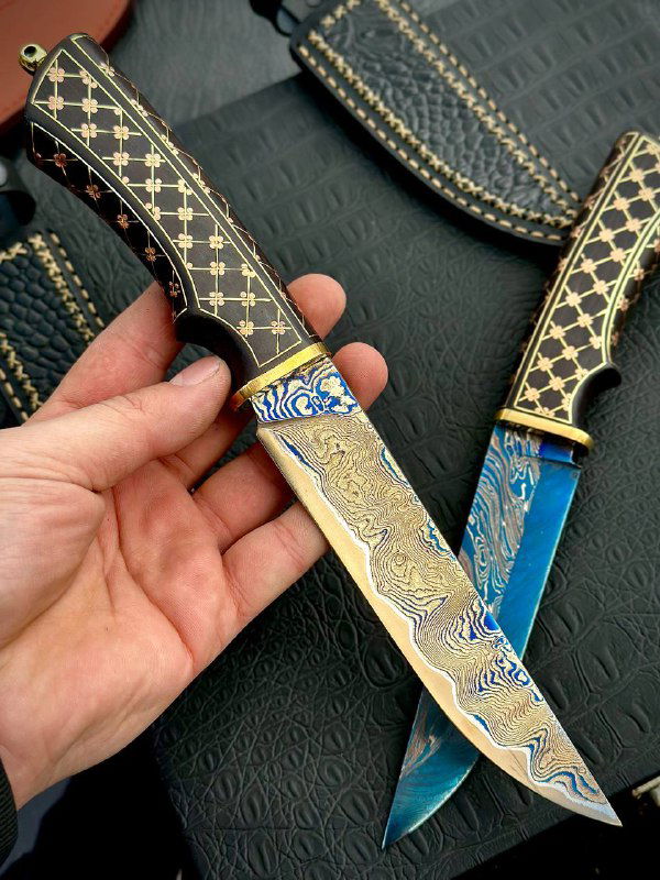 Ножи «Кобра» в руках мастера: виден золотистый и синий дамаск, латунные гарды и кожаные ножны на тёмном фоне.