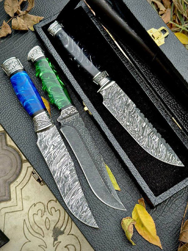 Ракурс, подчёркивающий форму клинка и отделку мельхиорового гарда; на фото три ножа с разной отделкой рукояти и кожаным футляром.