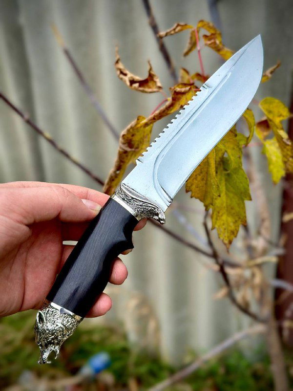 Вид клинка ножа «Волк» крупным планом: полированная рабочая кромка, зубчик на обухе и декоративная гарда на фоне листьев.