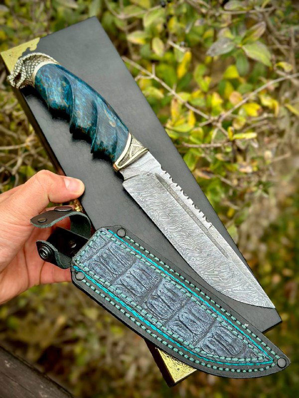 Фотография ножа «Кобра» и кожаных ножен на черной коробке; видно текстуру дамаска, отделку мельхиором и бирюзовую прострочку по ножнам.