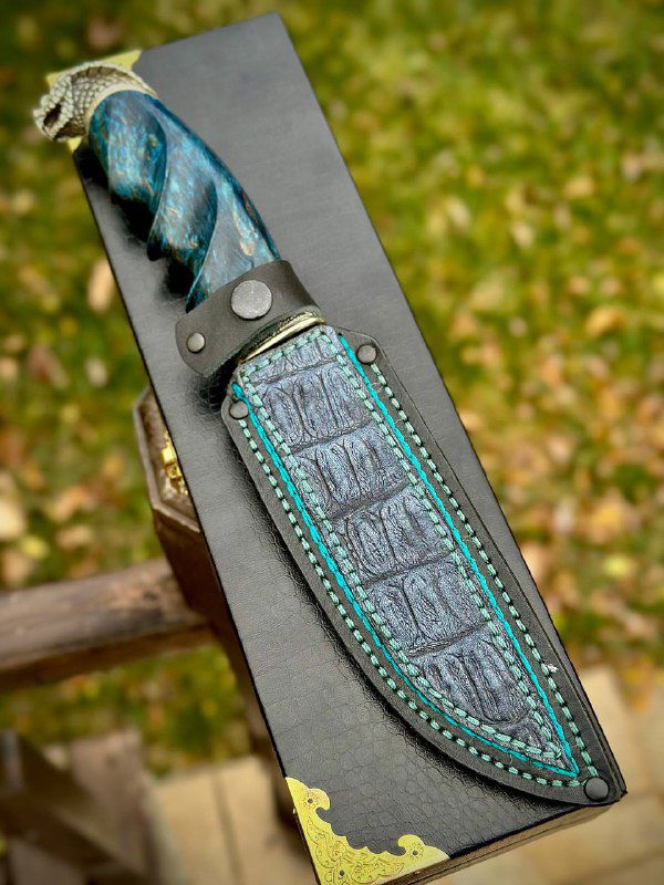 Ножны в комплекте: кожаные ножны с тиснением и бирюзовой прострочкой, лежащие рядом с ножом на деревянной поверхности и зеленью на заднем плане.