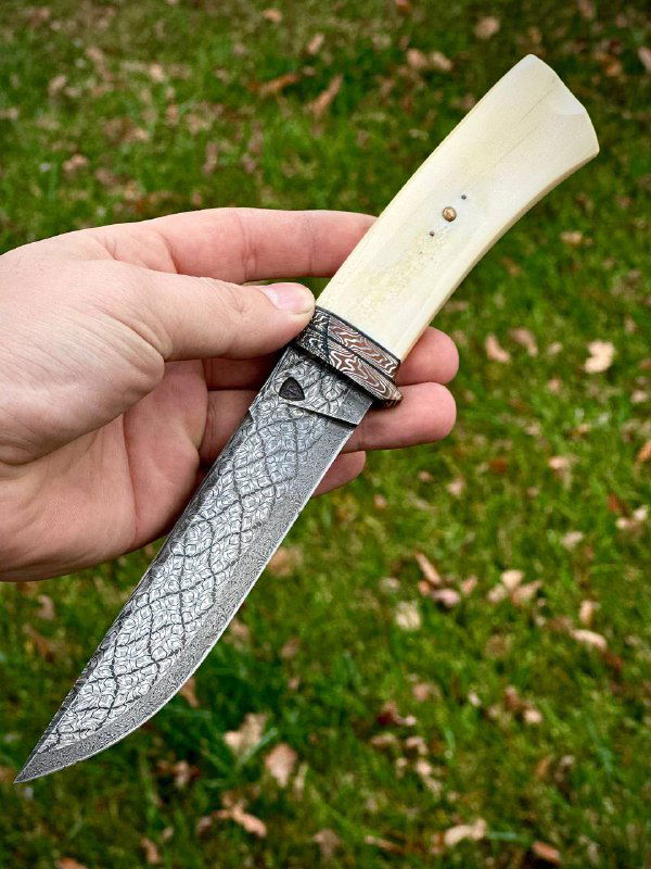 Рукоять ножа из клыка моржа в руке: вид крупным планом, видна ручная резьба и часть мозаичного дамаскового клинка на зелёном фоне.