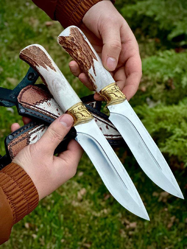 Две рукояти из рога лося и клинки ножей крупным планом, заметны латунные гарды и отражающая поверхность стали на фоне зелёного куста.