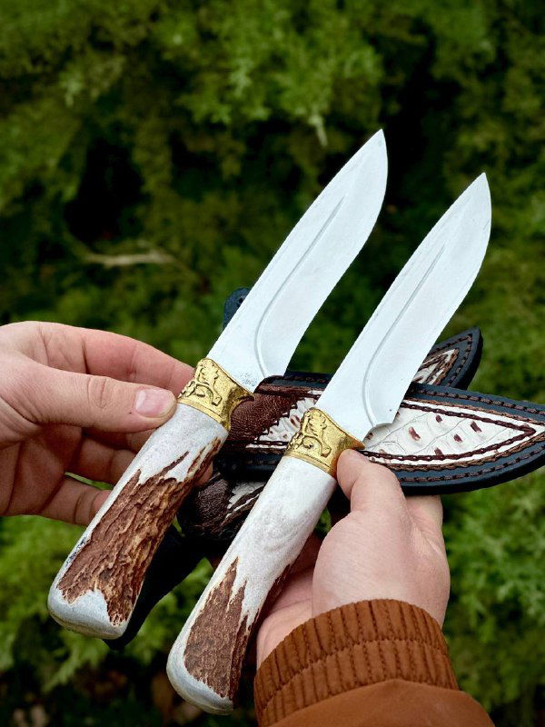 Крупный план двух клинков и рукоятей из рога лося: видно лезвие, текстуру рога и частично кожаные ножны на фоне кустарника.