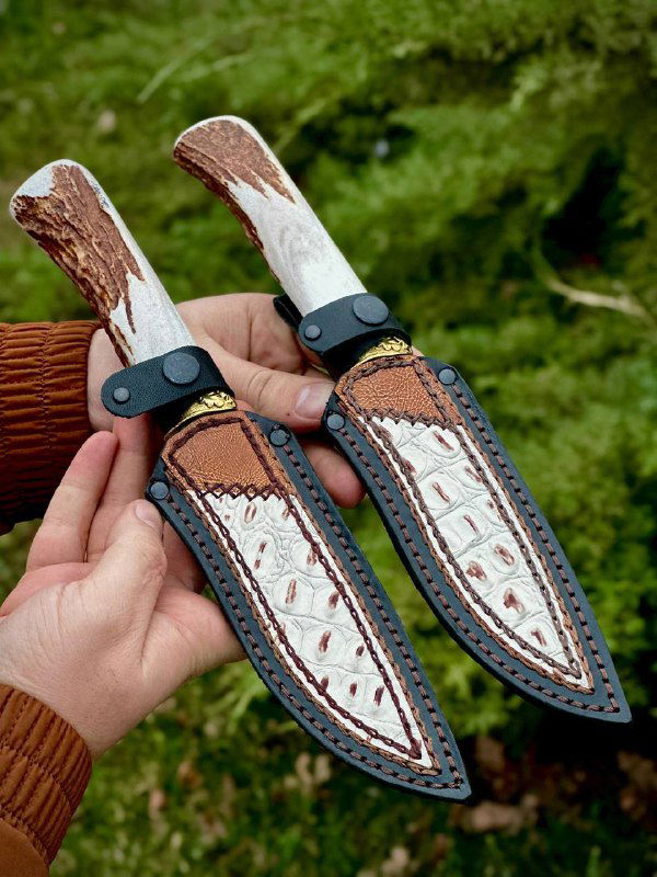 Руки держат два ножа в кожаных ножнах, показаны швы и декоративная отделка ножен, фон — зелёная растительность и кусты.