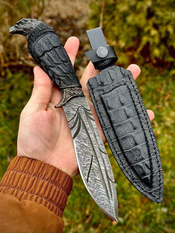 Нож «Ворон» с ножнами на ладони: видны мельхиоровые гарды, детальная резьба по дереву рукояти и кожаная кобура на природном фоне.