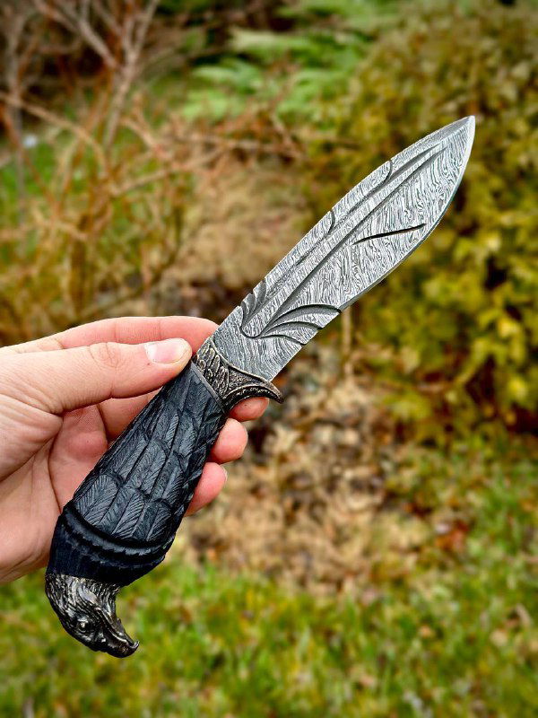 Вид клинка и рукояти ножа «Ворон» под углом: отчетливый рисунок дамасской стали, прорезная отделка и профиль лезвия, снято на открытом воздухе.