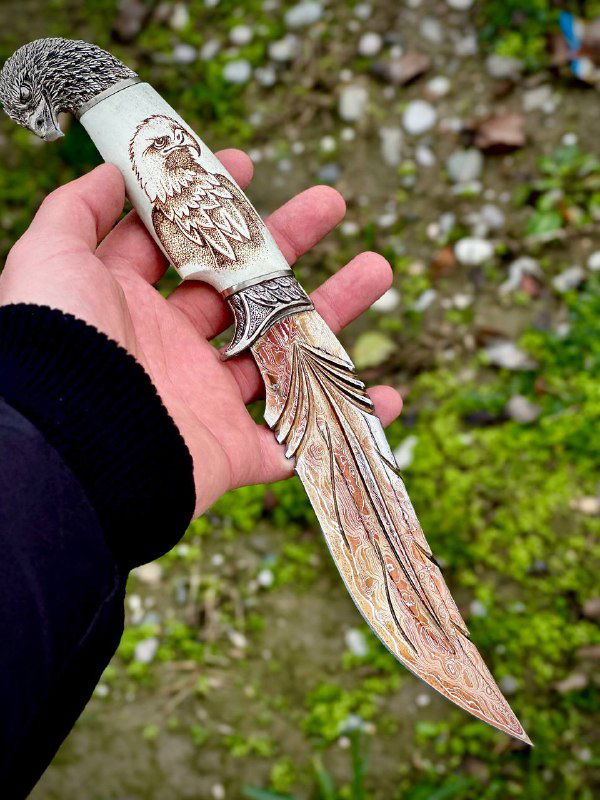 Крупный план второй рукояти ножа из рога лося с выжиганием орлана; фон — трава и земля, ручная съёмка на улице.