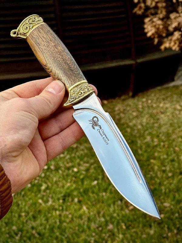 Рукоприёмное фото ножа «Скорпион»: нож держат в руке на фоне газона, видно отполированное лезвие и подпись на лезвии, рукоять из ореха с резными латунными элементами.