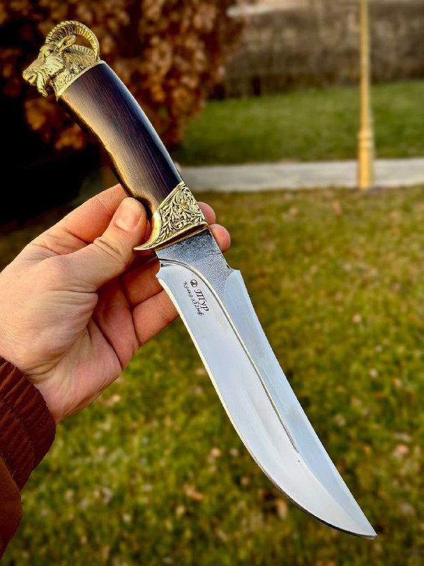 Крупный план ножа «Тур»: полированный клинок и детализированная латунная гарда, рукоять из граба и аккуратная отделка, фон — тот же уличный пейзаж.