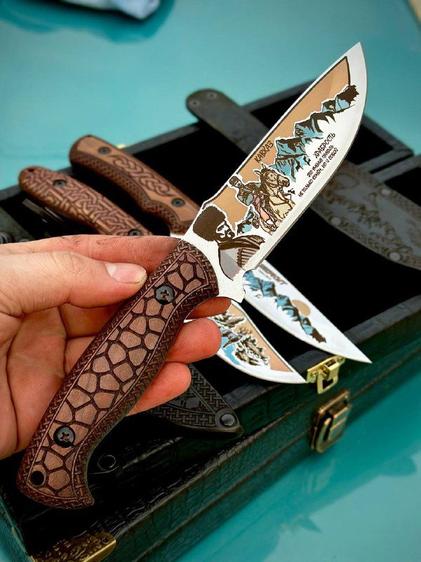 Рука держит разделочный нож с резной рукоятью из кавказского ореха и детализированной цветной гравировкой на лезвии, вид крупным планом.