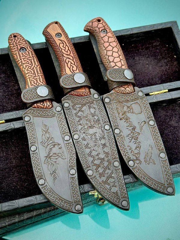 Три кожаные ножны с тисненым рисунком и матовой отделкой рядом с ножами; видно декоративную гравировку на ножнах и фурнитуре.
