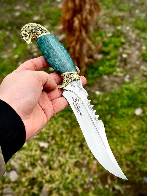 Рукоять зелёного ножа «Орлан» из карельской берёзы в руке мастера, видно ребро и зубчатую часть клинка из стали Х12мф и декоративную латунную гардy.
