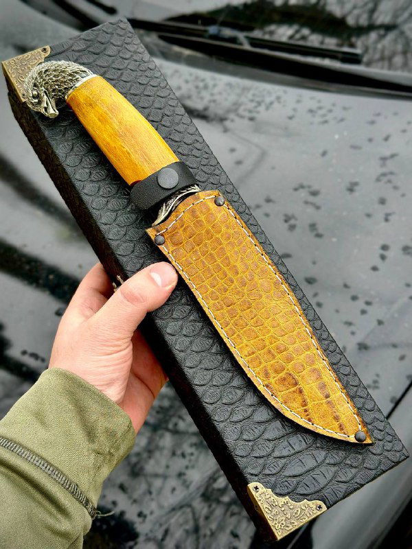 Рука держит нож на фоне мокрой чёрной поверхности; видна кожаная кобура жёлто-коричневого тона и чёткий узор дамасской стали на лезвии.